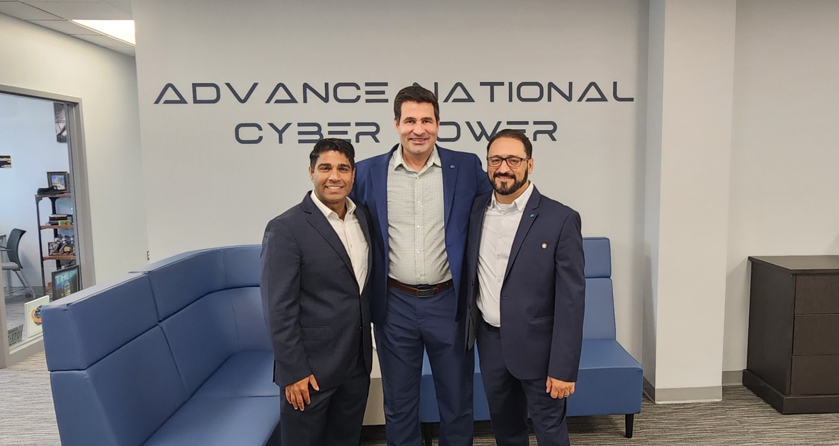 The image shows three men standing together indoors, all dressed in business attire — dark suits and light shirts — smiling for a professional photo. Behind them, a wall displays the words “ADVANCE NATIONAL CYBER POWER.” The setting appears to be a modern office or conference area, with a blue sectional couch, a dark wood cabinet, and a vase of blue flowers visible in the background. The overall atmosphere is professional and collegial, suggesting a formal visit, partnership, or meeting related to cybersecu