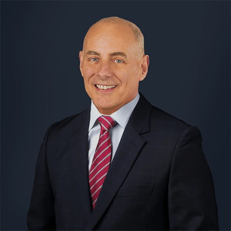 A person in a dark suit with a white shirt and red striped tie stands against a dark background.