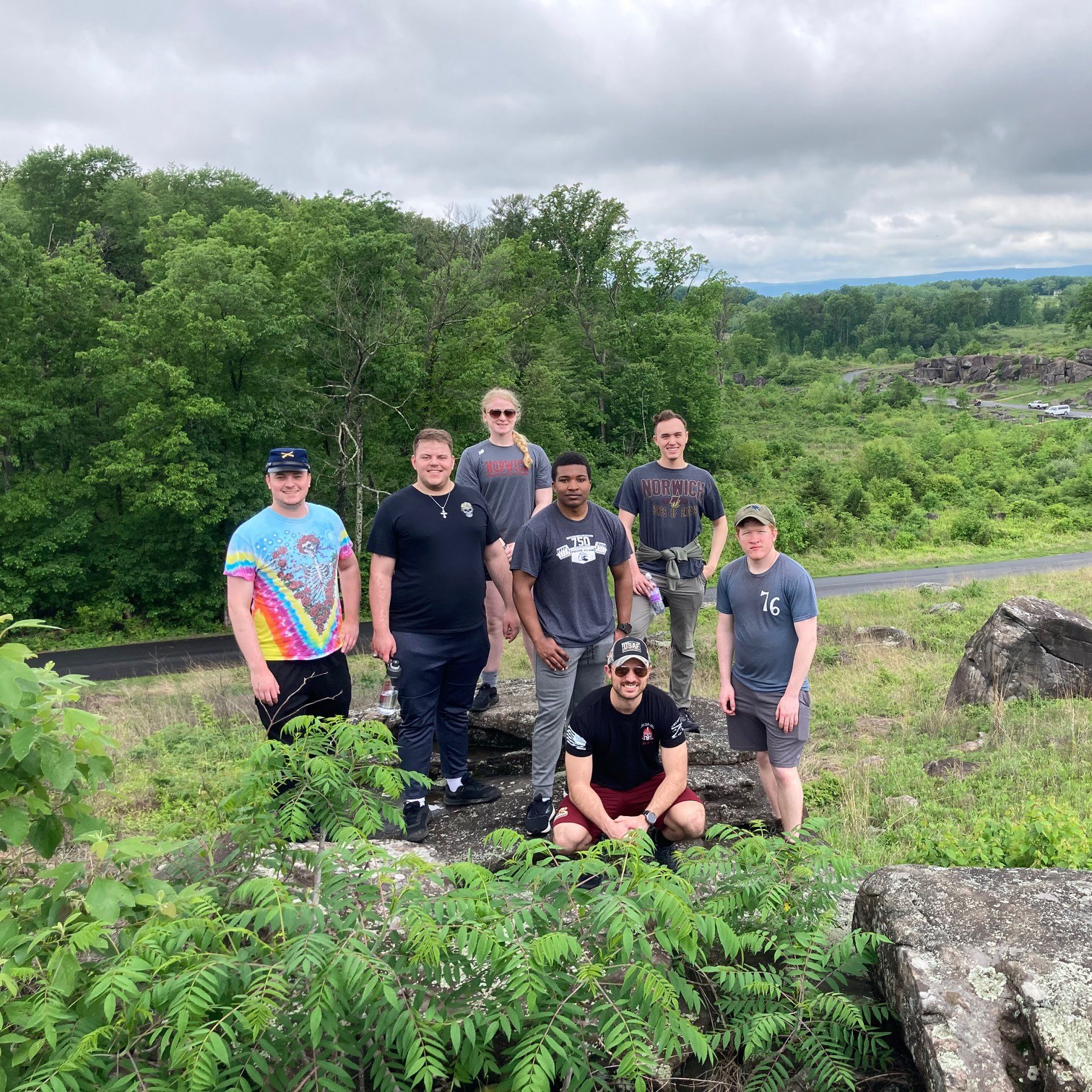 Seven people stand on a rocky hill with green fields, trees, and a winding road in the background.