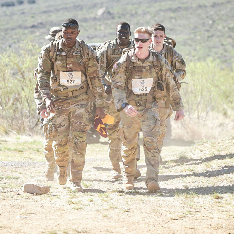Cadets in uniform with backpacks and race bibs walk on a dirt trail.