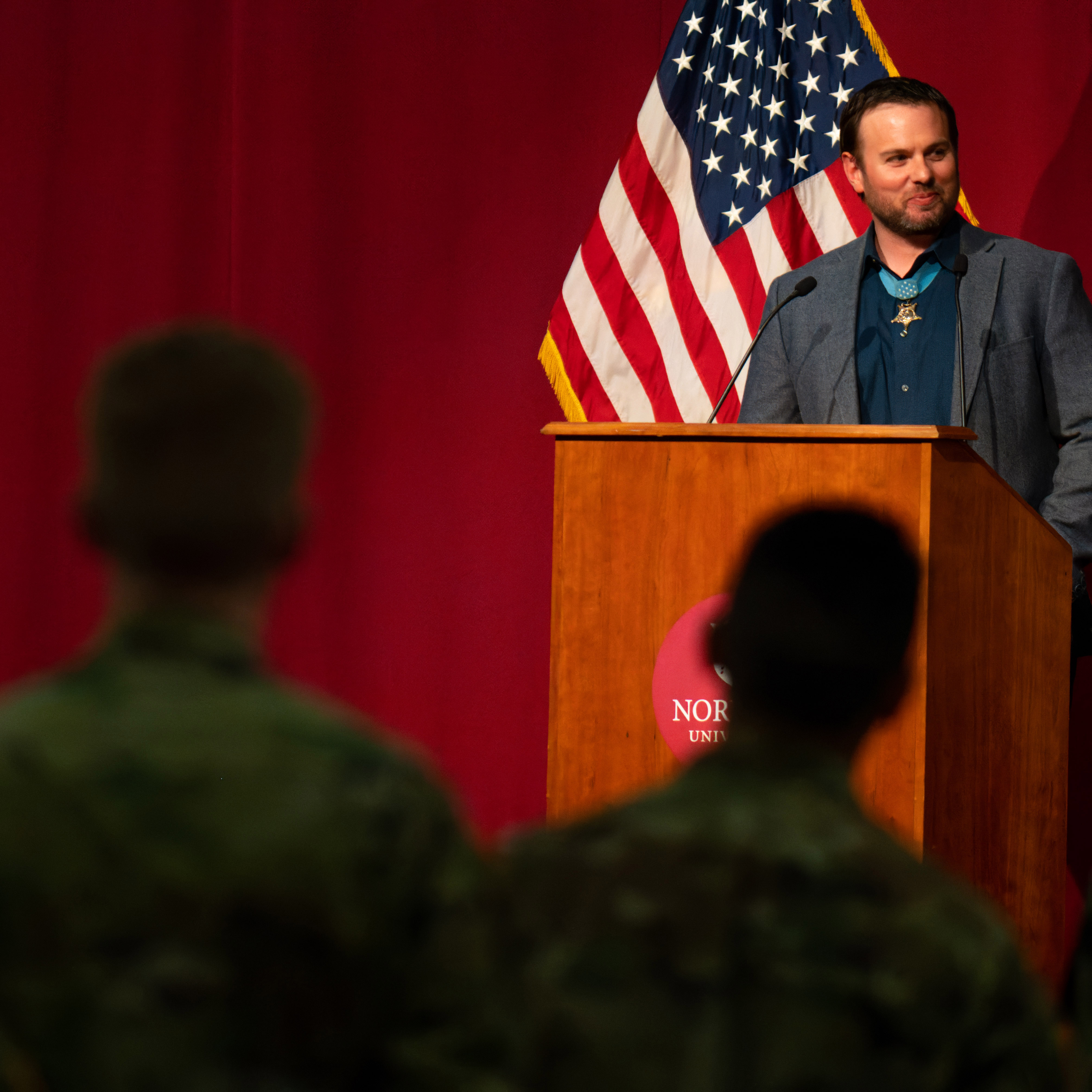 Speaker stands at a Norwich University podium with an American flag behind him.