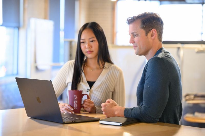 Two people sit at a table looking at a laptop, with one holding a Norwich mug.