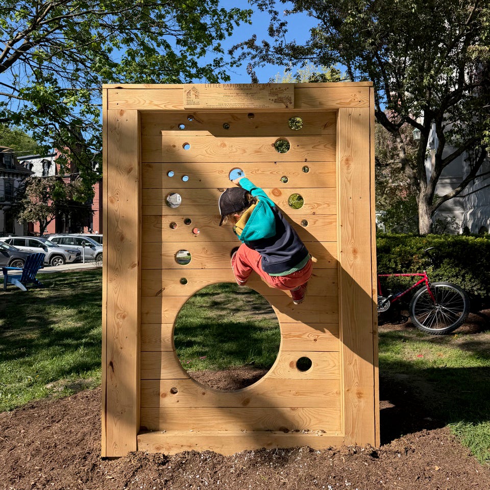 Child climbs a wooden panel with circular openings in an outdoor play area.