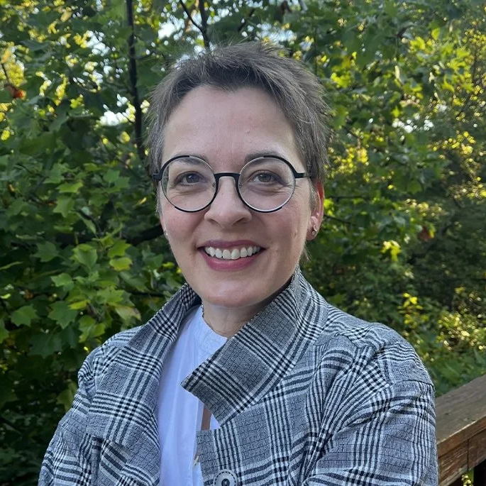 Person with short hair and round glasses smiles outdoors in front of leafy trees while wearing a plaid jacket.