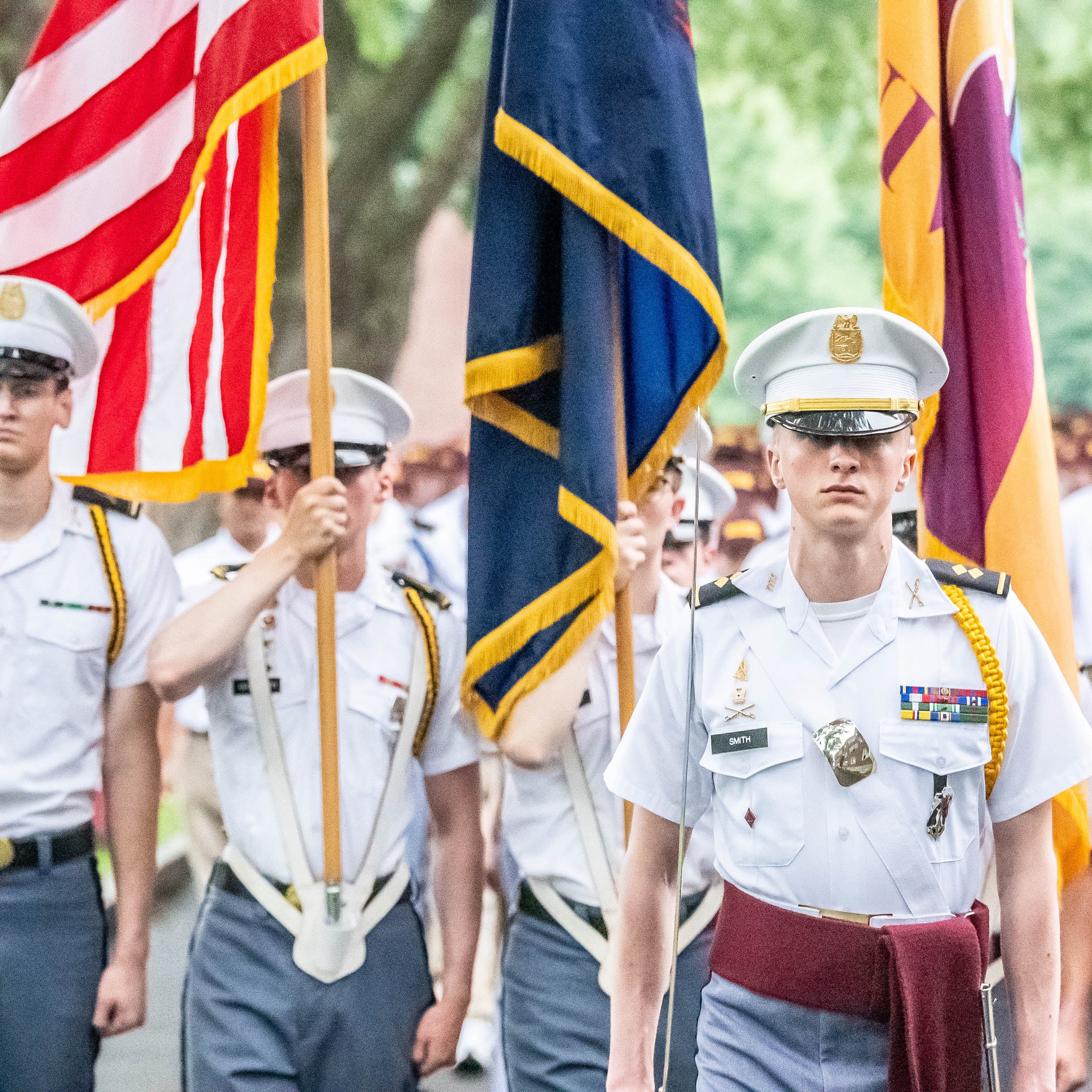 Cadets in uniform march in formation carrying the American flag and ceremonial flags during a parade.