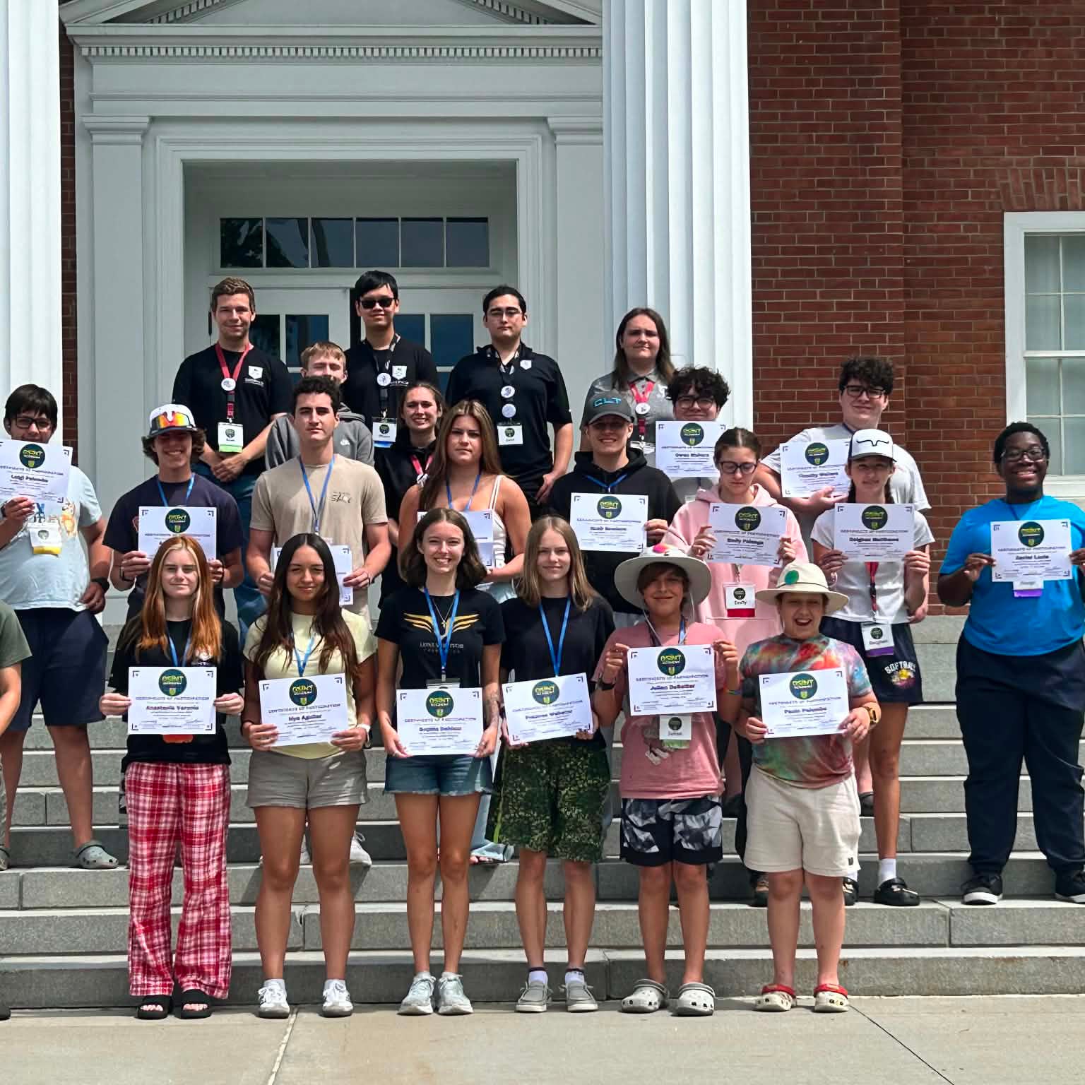 Group of students stand on building steps holding certificates in front of a brick building with white columns.