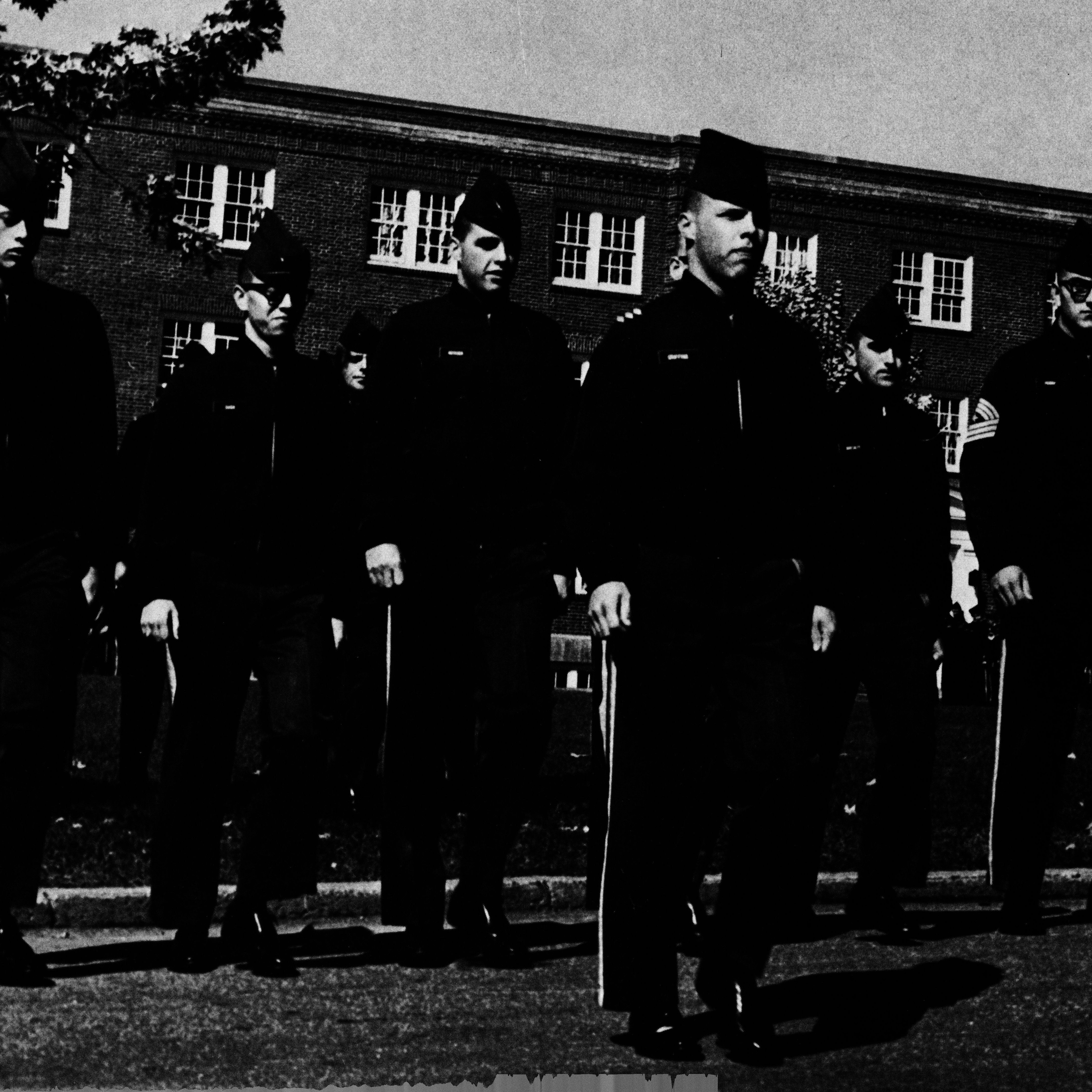 Cadets in uniform march in formation outside a brick building.