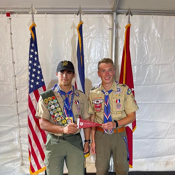 Two scouts in uniform hold a Norwich University pennant in front of three flags.