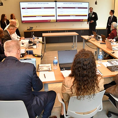 Presenter speaks to attendees seated around tables as two screens display a legacy fleet presentation.