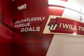 A Norwich University hallway features bold crimson and gold wall graphics. On the left, the NU logo is paired with the motivational phrase “Relentlessly pursue all of your goals” over a faint image of a helicopter. To the right, another NU logo accompanies the school’s motto, “I Will Try,” mounted on a curved sign.