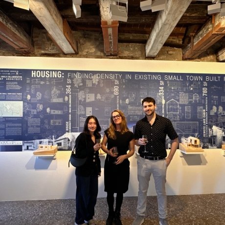 Three individuals holding drinks stand in front of an exhibition panel titled "HOUSING: FINDING DENSITY IN EXISTING SMALL TOWN BUILT FABRIC" at an architectural gallery. They are smiling.