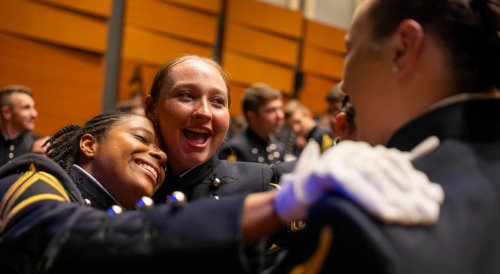 Three cadets in formal military attire greet each other at an event in an auditorium.