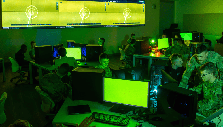 Students in military uniforms working at desktop computers in a green-lit Norwich University cybersecurity lab, with digital displays showing network diagrams.