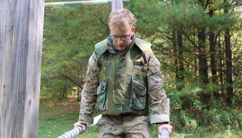 A person in military camouflage uniform performing an exercise on parallel bars in an outdoor obstacle course. The background features a wooded area with green foliage.