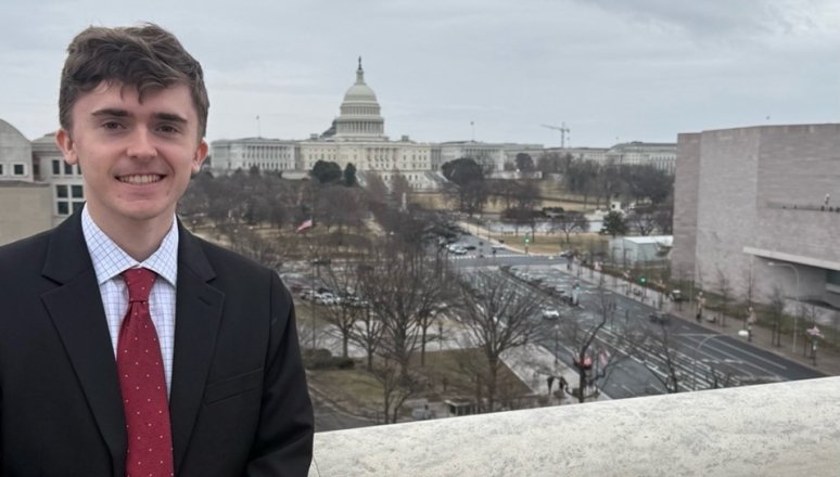 A student in a black suit stands on the Speaker's Balcony with D.C. behind him.