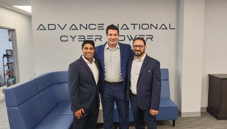 The image shows three men standing together indoors, all dressed in business attire — dark suits and light shirts — smiling for a professional photo. Behind them, a wall displays the words “ADVANCE NATIONAL CYBER POWER.” The setting appears to be a modern office or conference area, with a blue sectional couch, a dark wood cabinet, and a vase of blue flowers visible in the background. The overall atmosphere is professional and collegial, suggesting a formal visit, partnership, or meeting related to cybersecu