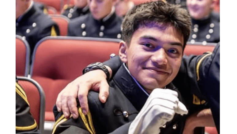 A cadet in a formal military uniform sits in an auditorium seat, smiling at the camera while raising a gloved hand in a friendly gesture