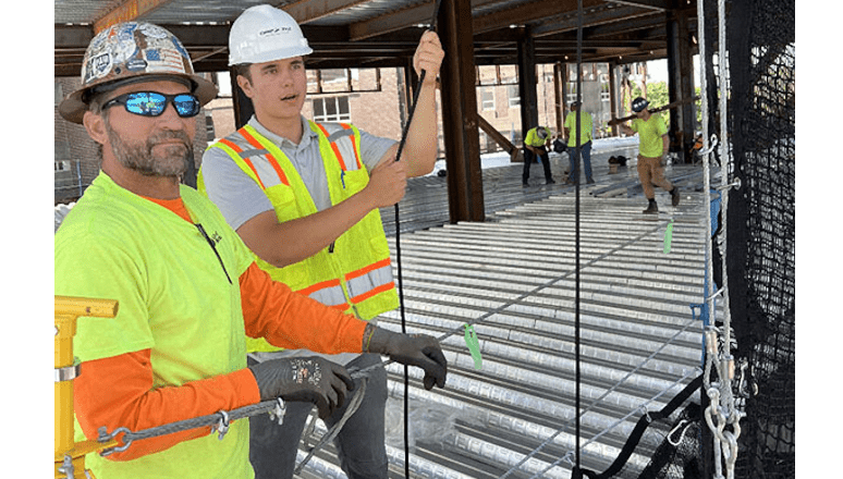wo construction workers wearing hard hats and safety vests work with cables on a steel building frame, while others work in the background.