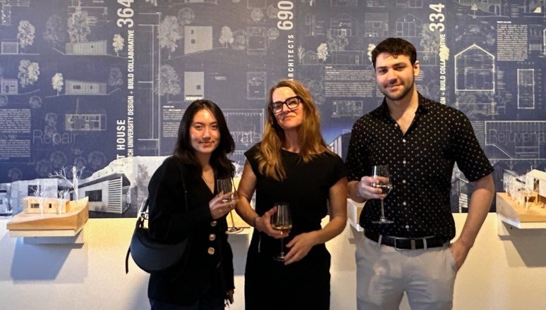 Three individuals holding drinks stand in front of an exhibition panel at an architectural gallery. They are smiling.