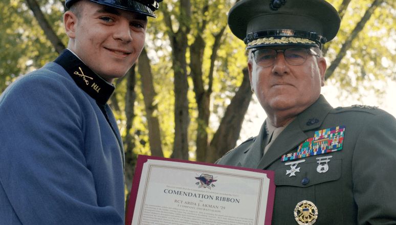 Two uniformed service members stand outdoors holding a commendation certificate together.