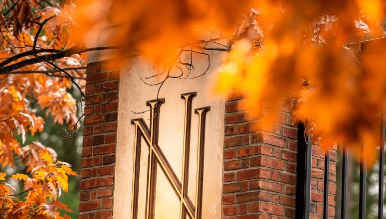 A brick pillar with the logo "NU" on a gold plaque, framed by vibrant orange autumn leaves.
