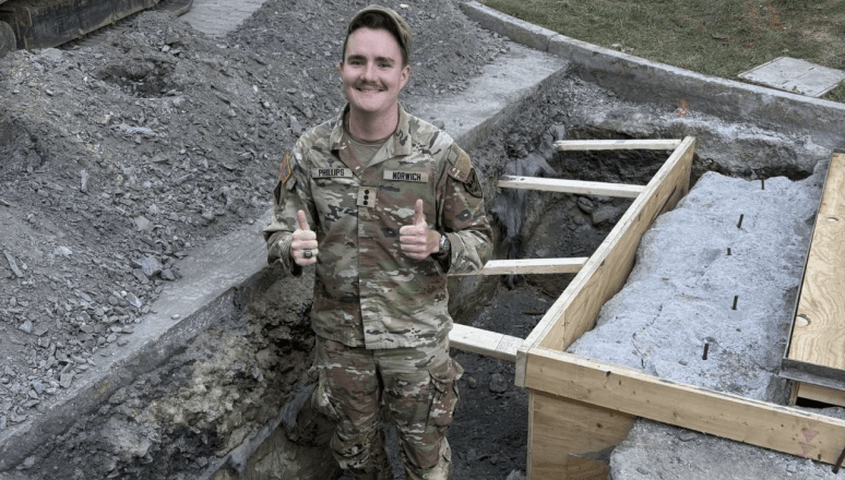 A person in military uniform stands in a shallow construction trench giving two thumbs up, with gravel piles, wooden forms, and excavation equipment nearby.