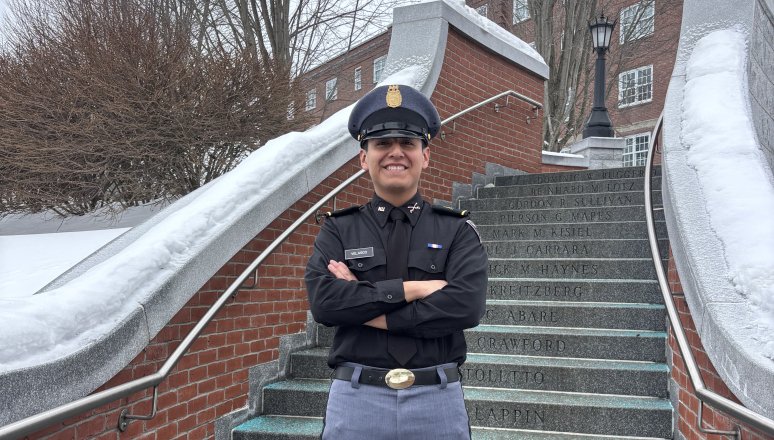 Person in a dark uniform and cap stands with arms crossed on outdoor stone steps bordered by brick walls and snow.