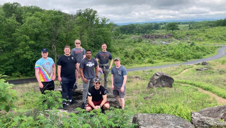 Seven people stand on a rocky hill with green fields, trees, and a winding road in the background.