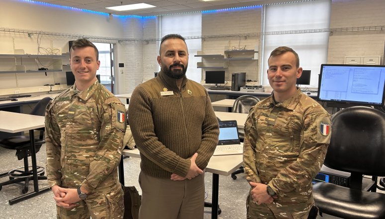 Two cadets and an instructor stand in a computer lab classroom.