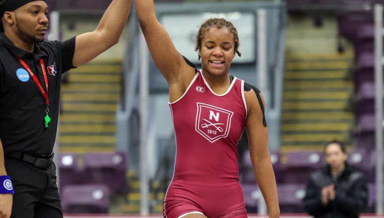 A wrestler in a maroon uniform stands on a mat as a referee lifts one arm in victory, with empty seats and a few people in the background.