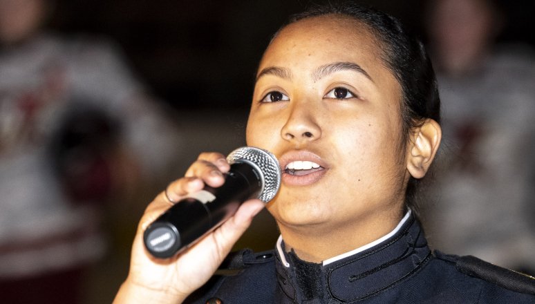 Cadet in dress uniform sings into a microphone indoors.
