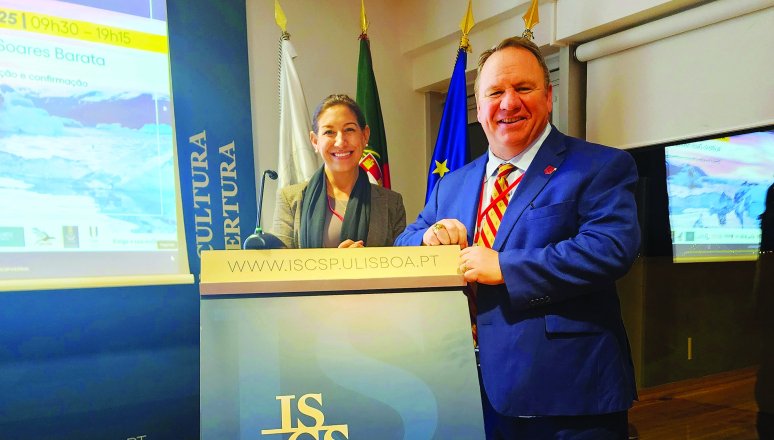 Two people stand at an ISCTE podium in a conference room with flags and presentation screens behind them.