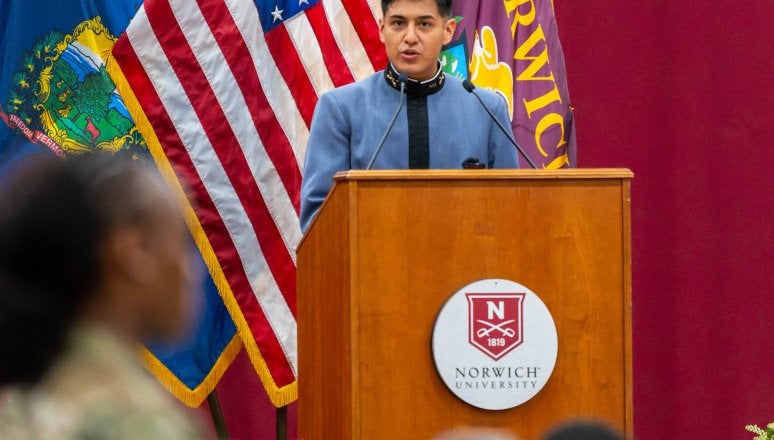 Cadet speaks at a Norwich University podium with American, Vermont, and Norwich flags behind.