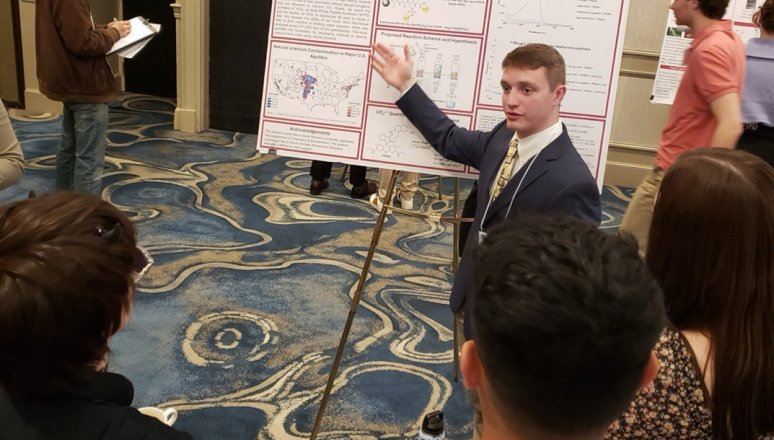 Student presents a research poster to a group at an indoor academic conference.