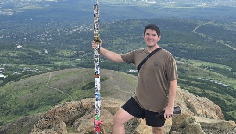 Person stands on rocky summit holding a pole with an American flag above a valley and distant water.