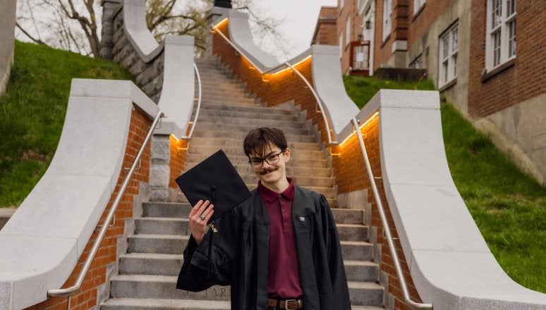 Graduate holds cap while standing on campus stairs between brick buildings.