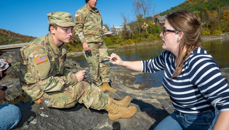 Instructor shows measuring tool to cadets seated on rocks beside a river during outdoor fieldwork.