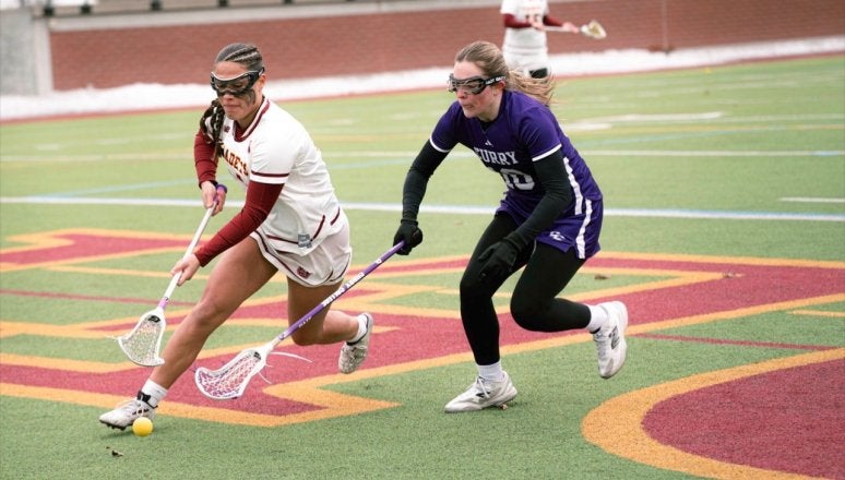 Norwich lacrosse player carries the ball while a Curry defender runs beside them on the field.