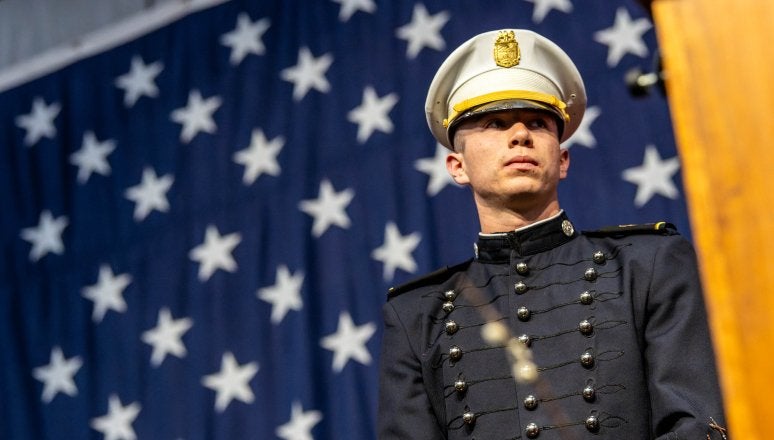 Cadet in dress uniform stands before large American flag during ceremony.