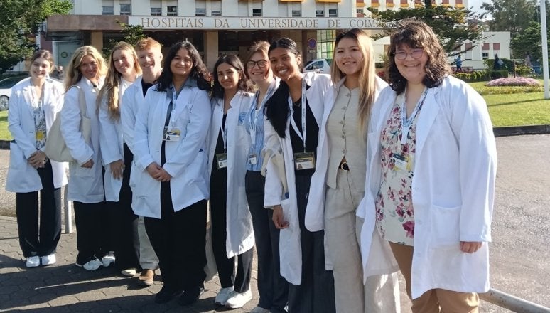 Group of students in white lab coats stand outside Coimbra University Hospitals building.