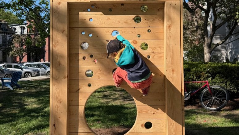 Child climbs a wooden panel with circular openings in an outdoor play area.