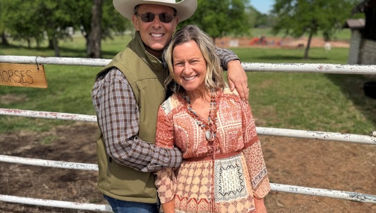 Two people stand together in front of a ranch gate, with one wearing a cowboy hat and both smiling at the camera.