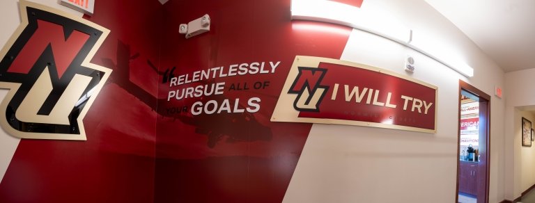 A Norwich University hallway features bold crimson and gold wall graphics. On the left, the NU logo is paired with the motivational phrase “Relentlessly pursue all of your goals” over a faint image of a helicopter. To the right, another NU logo accompanies the school’s motto, “I Will Try,” mounted on a curved sign.
